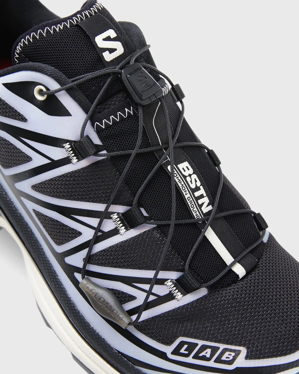 Close detail of bungee laces, BSTN Common Ground tag, S logo, LAB badge, lilac overlays, 5 AM