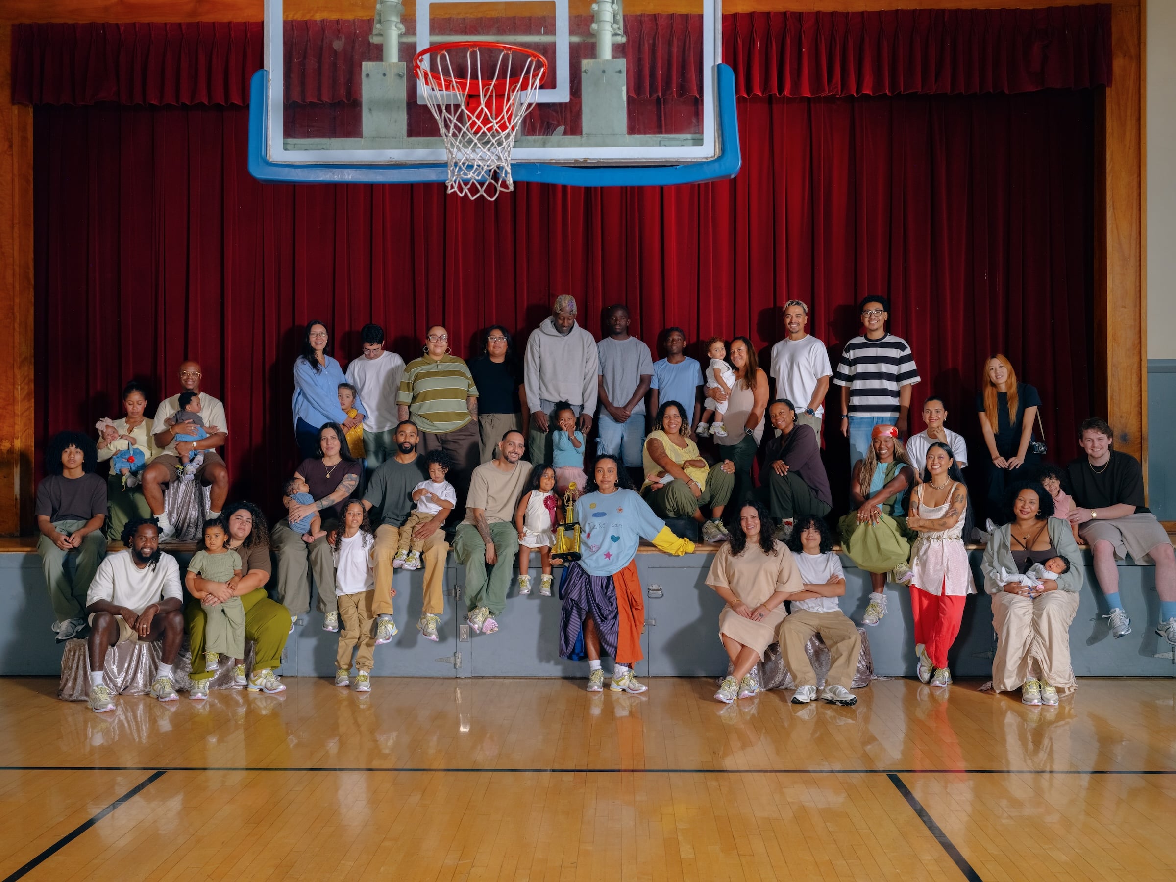 Large group posed on a gym stage in front of red curtains and a basketball hoop, many wearing similar sneakers, x Evelynn