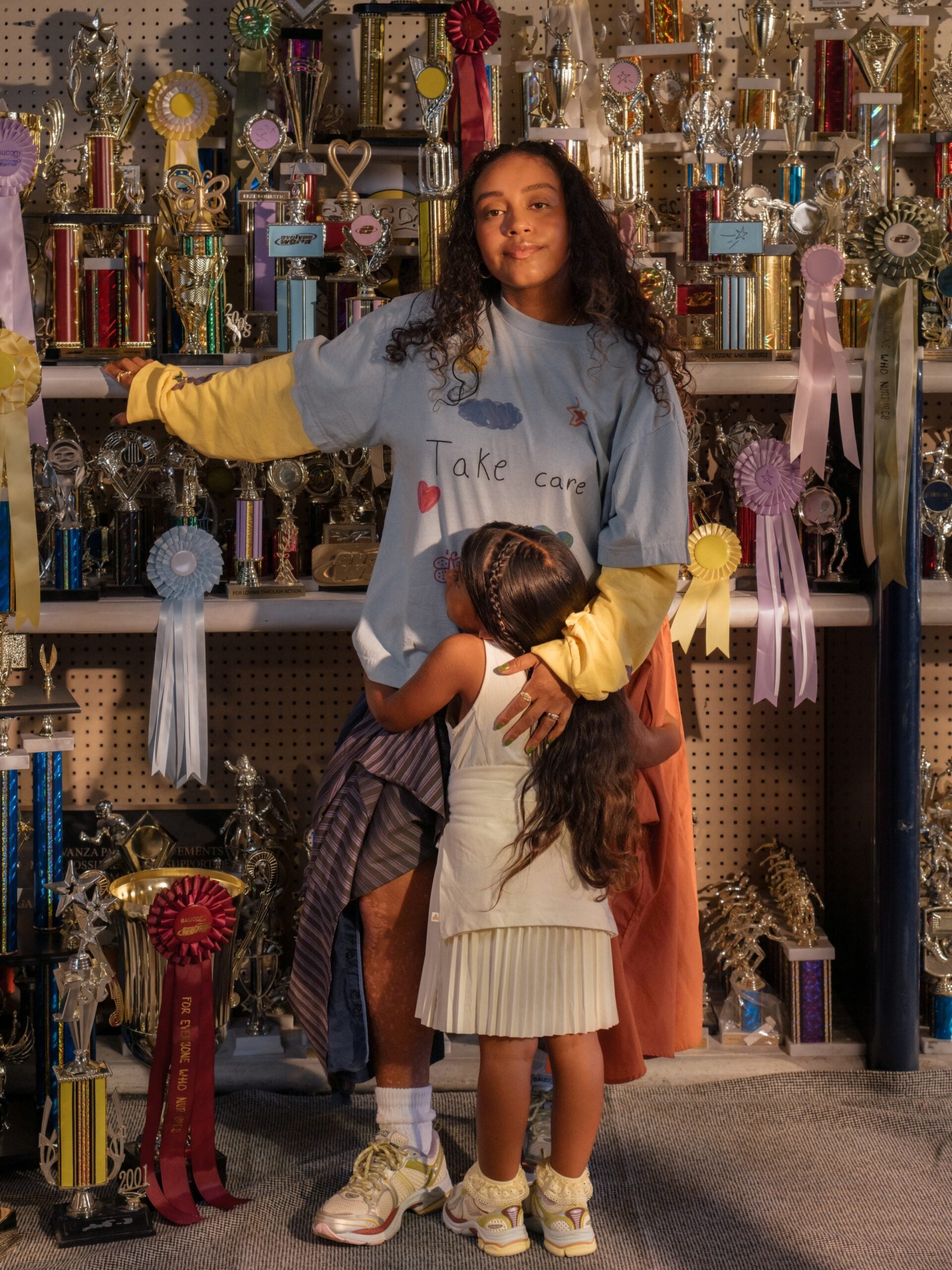 Person in a Take care shirt hugged by a child, in front of shelves of trophies and ribbons, both wearing sneakers, x Evelynn