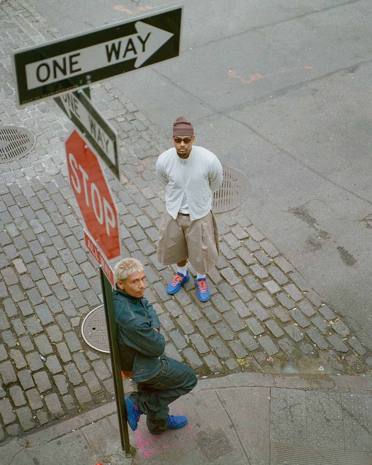High-angle street shot of two men in blue sneakers by One Way and Stop signs on cobblestones, NYC Blue