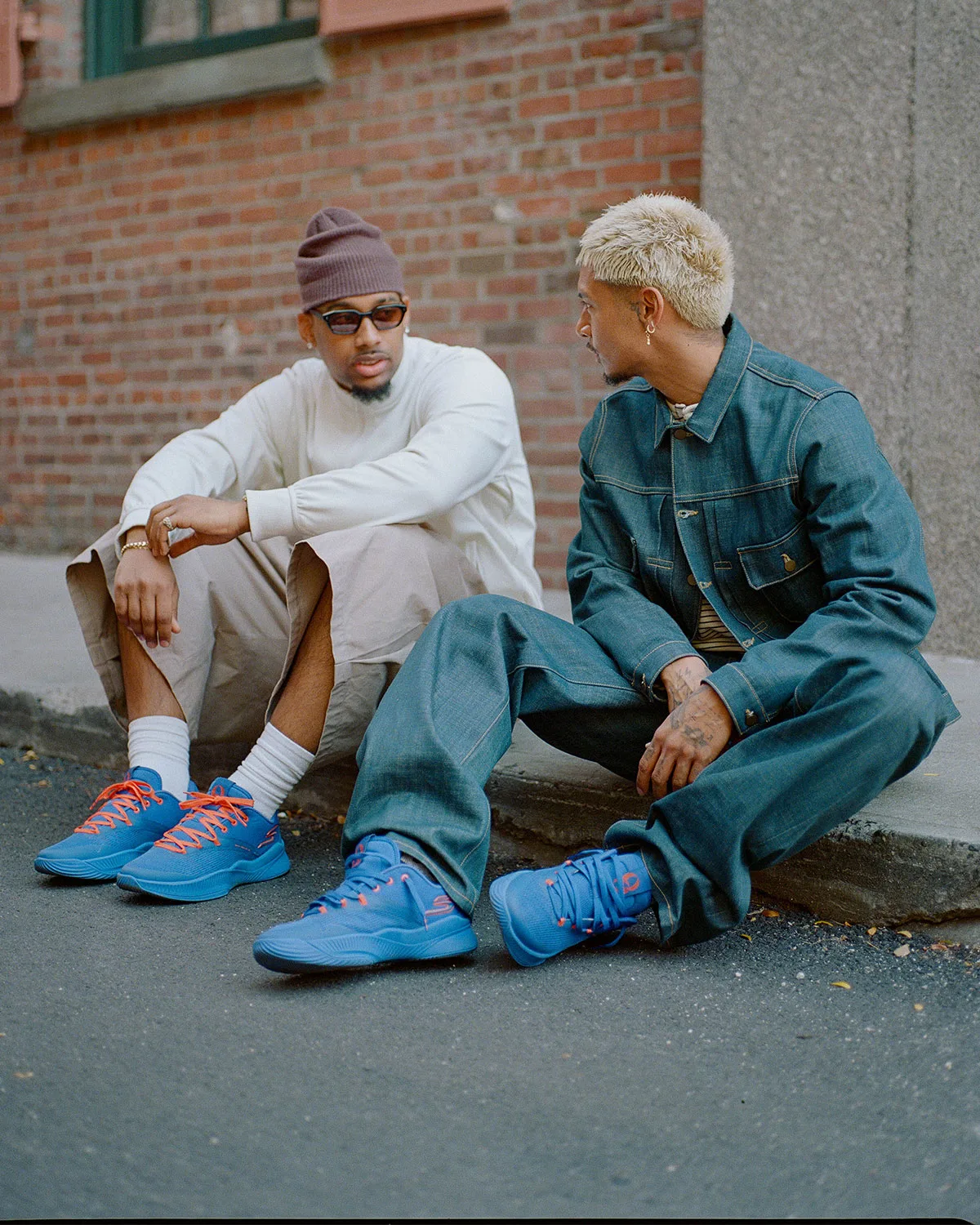 On-foot shot of two seated men in blue sneakers with orange laces, brick wall and curb behind, NYC Blue