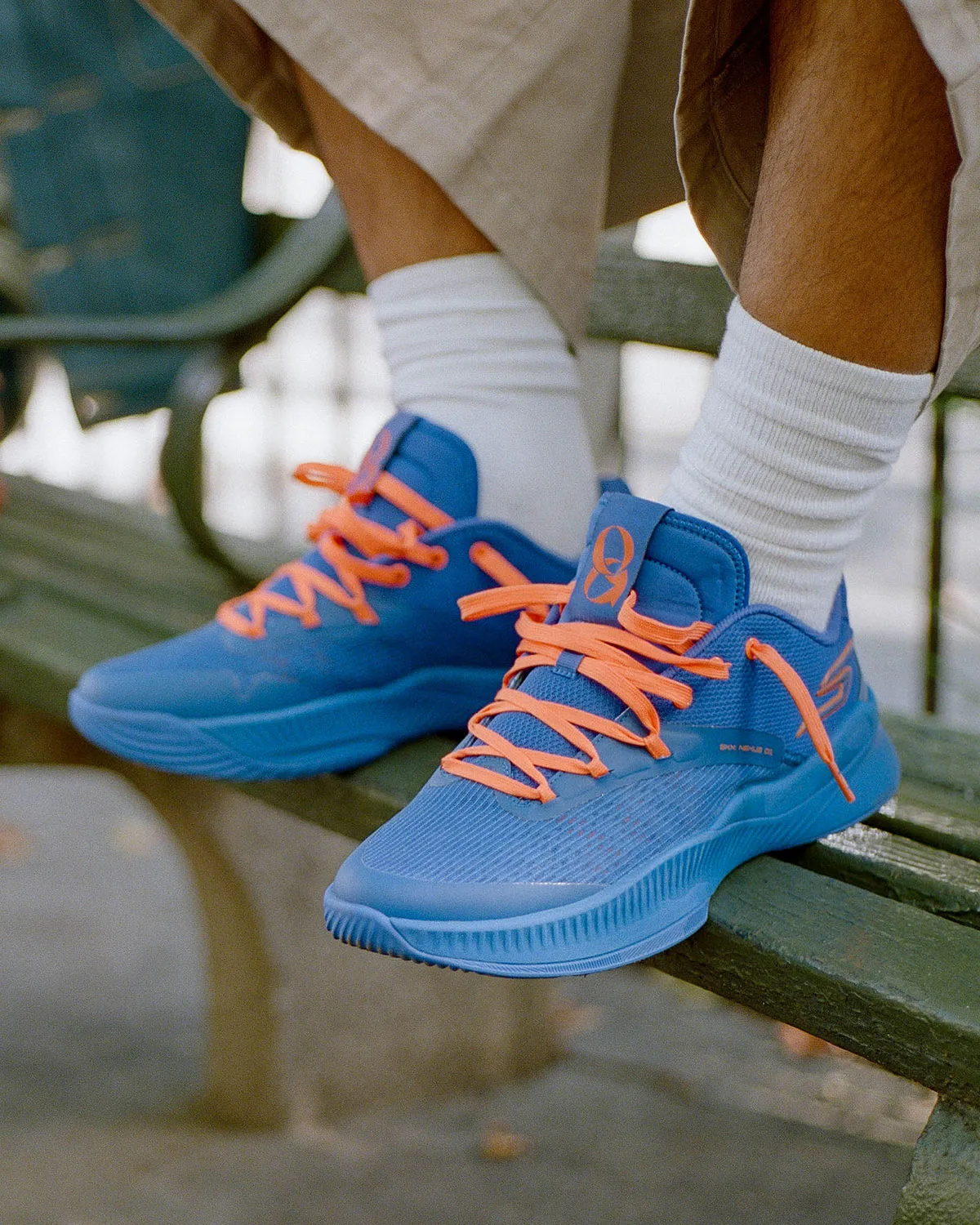On-foot view on a bench, blue mesh upper, orange laces, white crew socks, orange logo on heel tab, NYC Blue