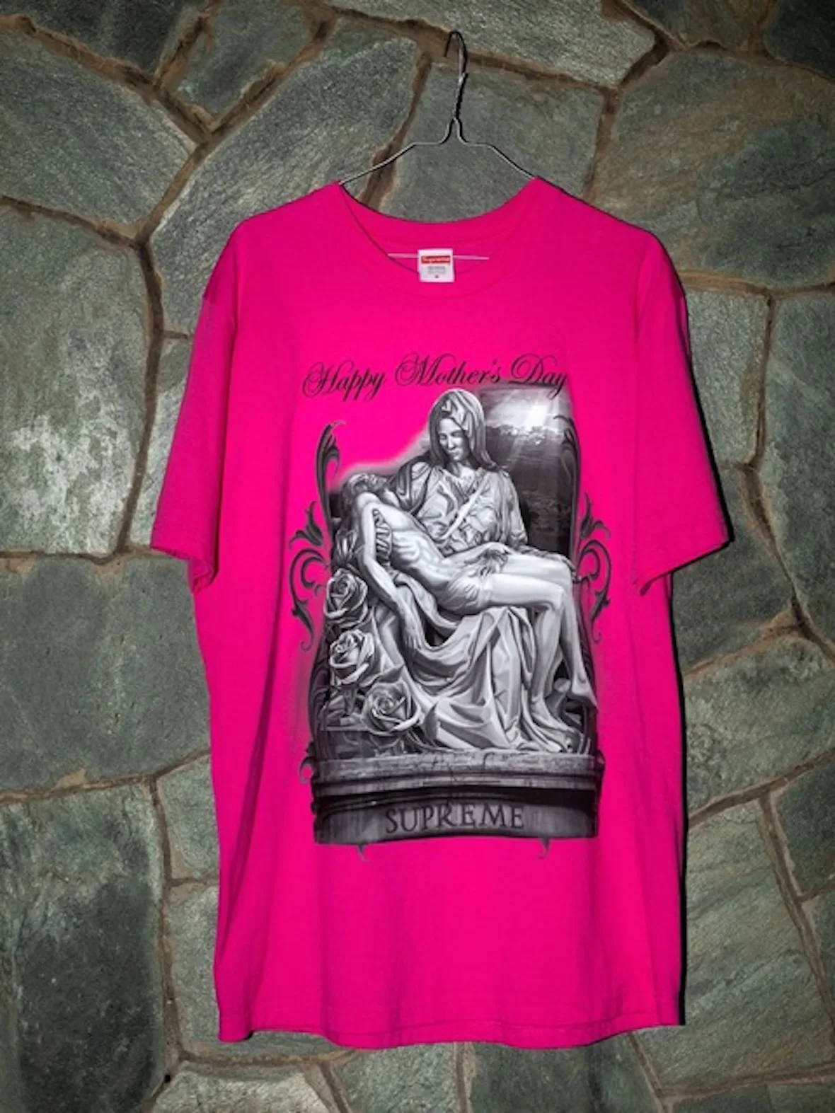Bright pink tee on a hanger against stone wall, with grayscale pietà graphic and Mother's Day text, 2026 Tees Collection