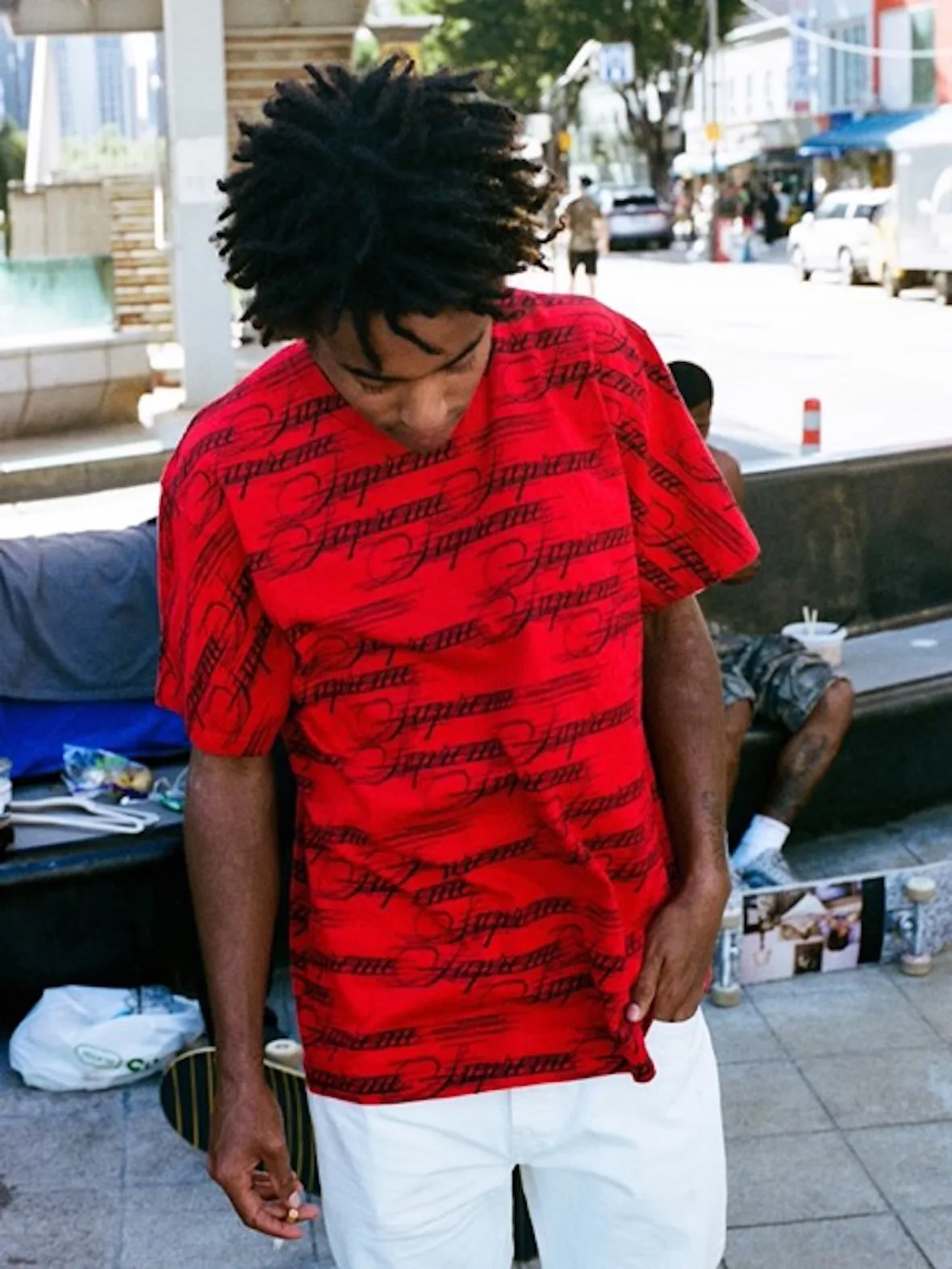 On-foot street shot of person in red tee with black script Supreme pattern, skateboards behind, 2026 Tees Collection