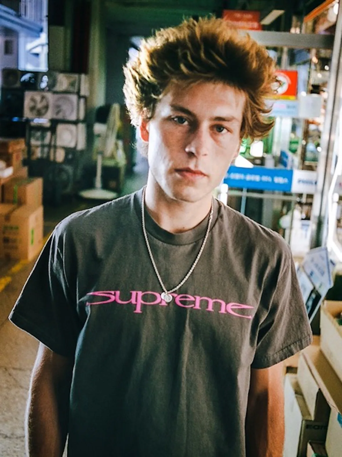 On-foot shot of person in grey tee with pink Supreme logo and silver chain, in a busy market aisle, 2026 Tees Collection