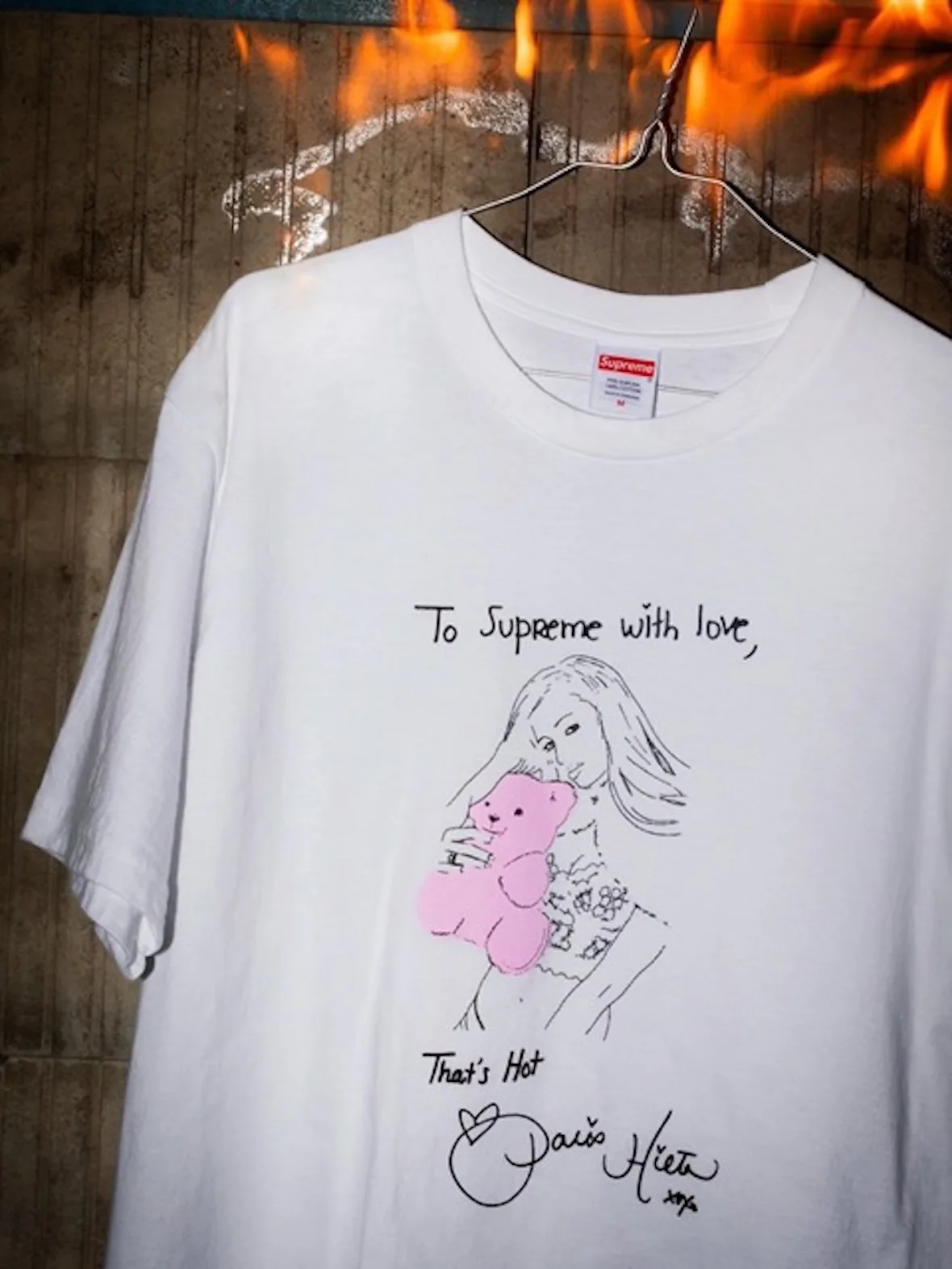 Close-up of white tee on a wire hanger with pink teddy bear drawing, flames behind, 2026 Tees Collection
