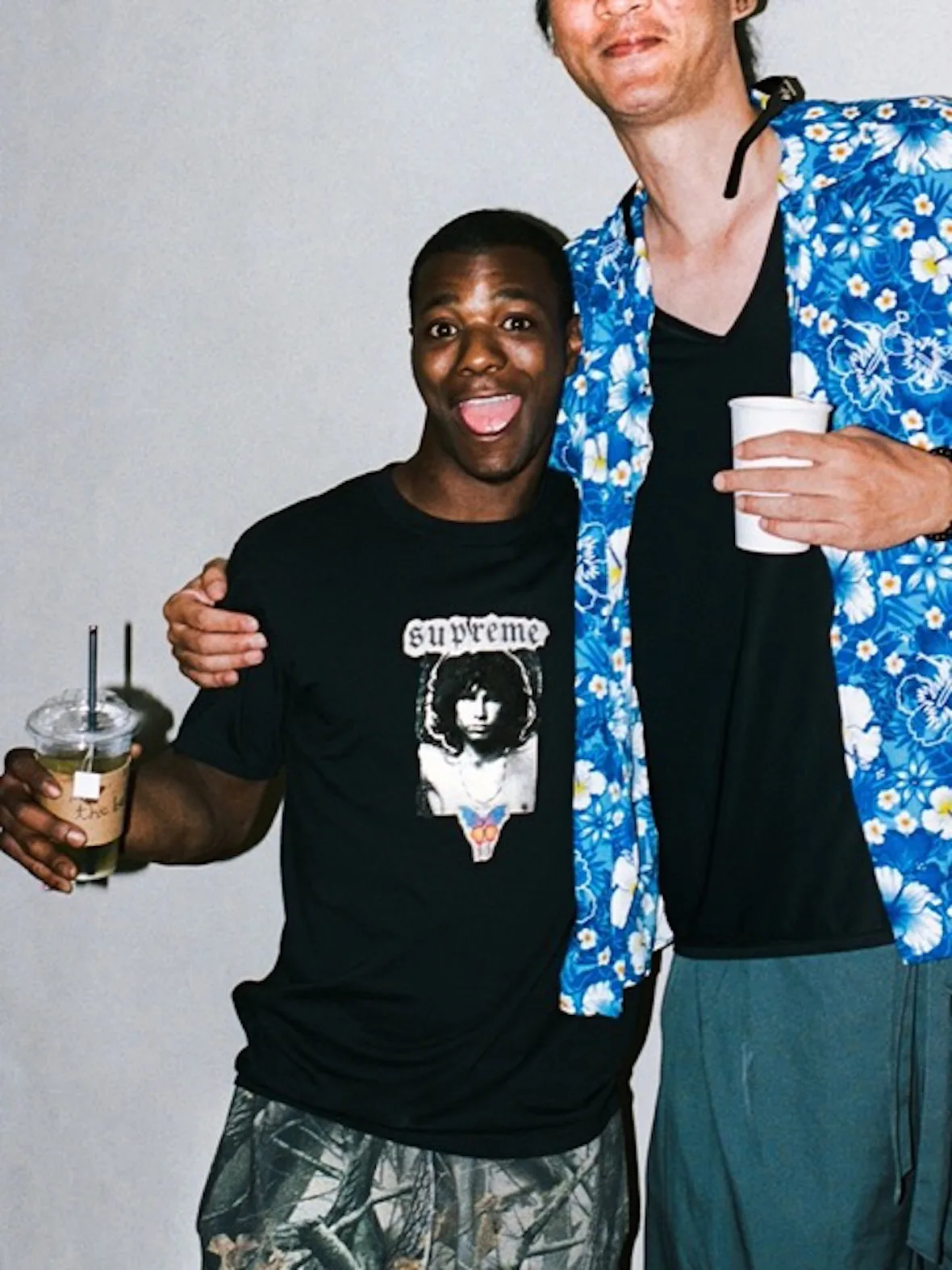 Two people posing, one in black tee with Supreme portrait graphic, other in blue floral shirt, 2026 Tees Collection