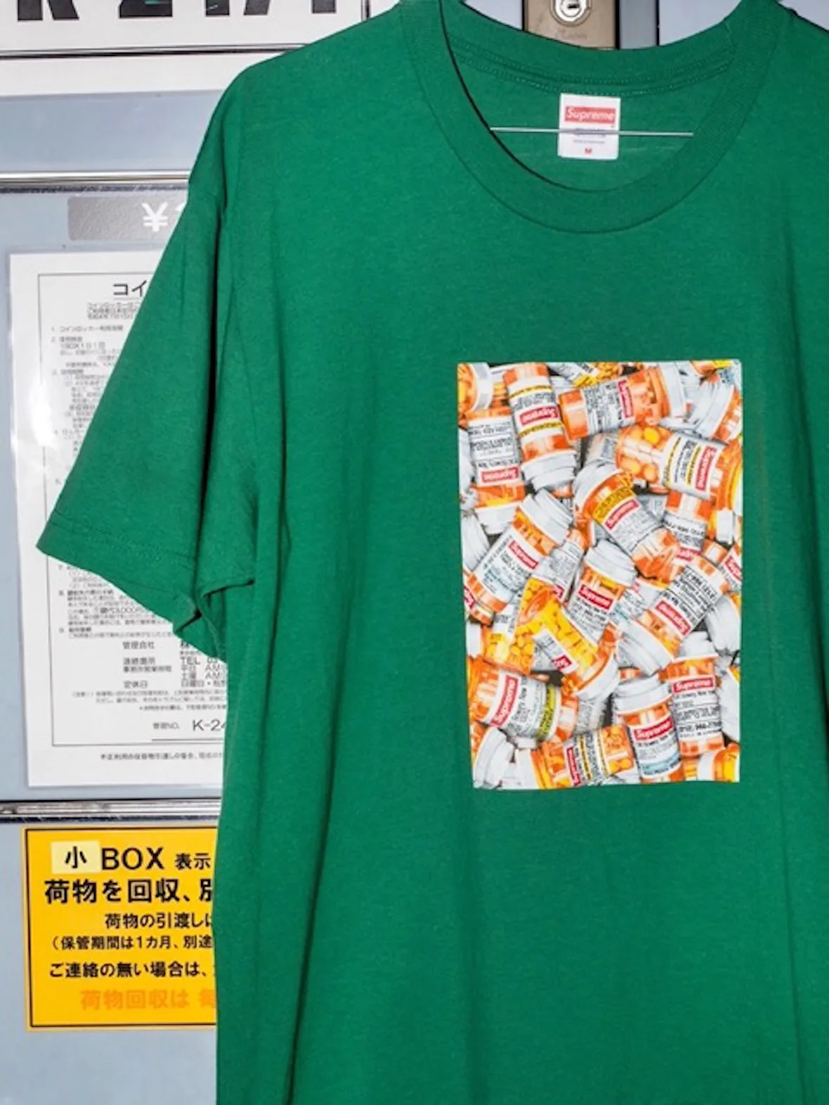 Close-up of green tee with pill bottle print, hanging in front of Japanese signage, 2026 Tees Collection