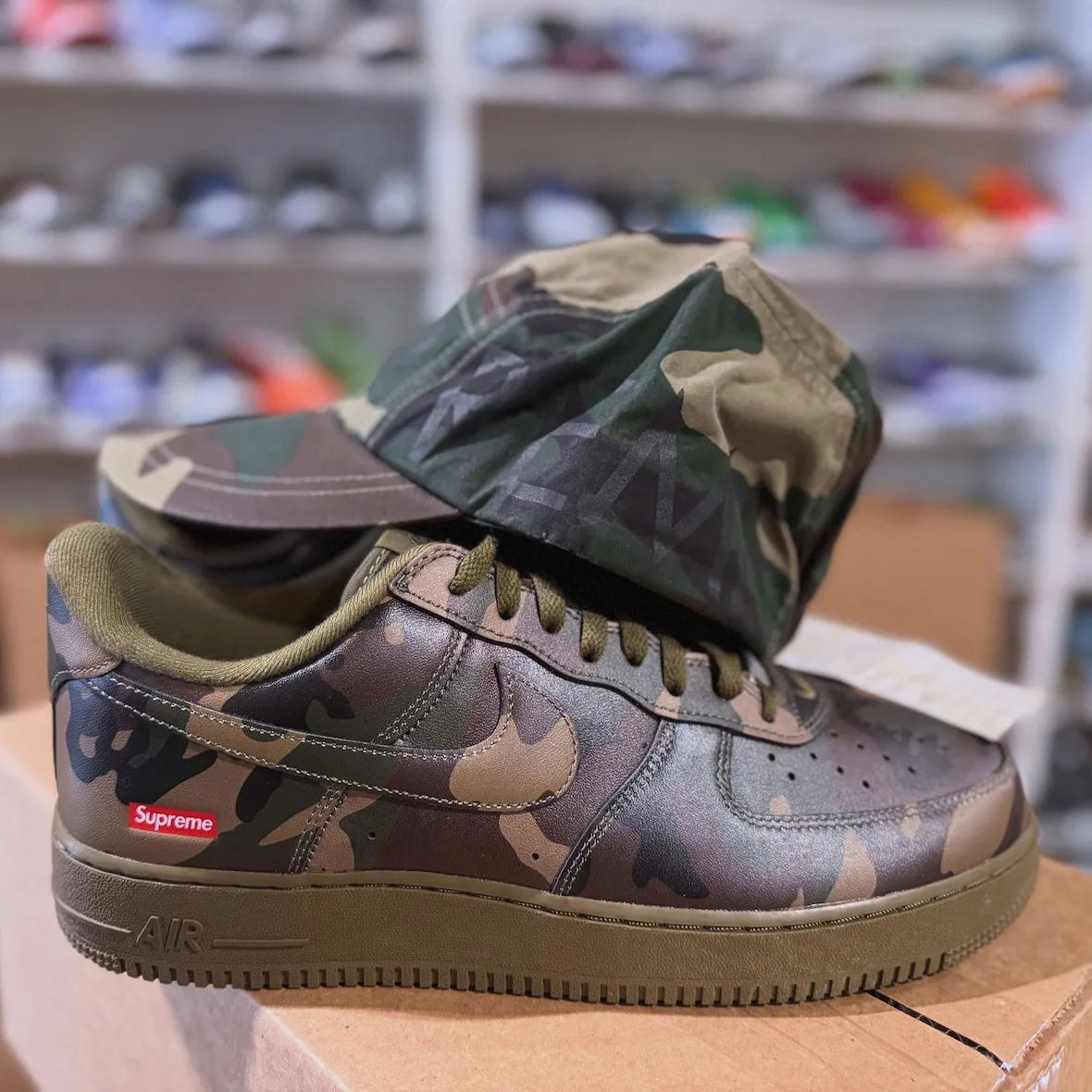 Lateral close view of woodland camo sneaker, olive laces and sole, camo cap above, shoe shelves behind, Olive Camo