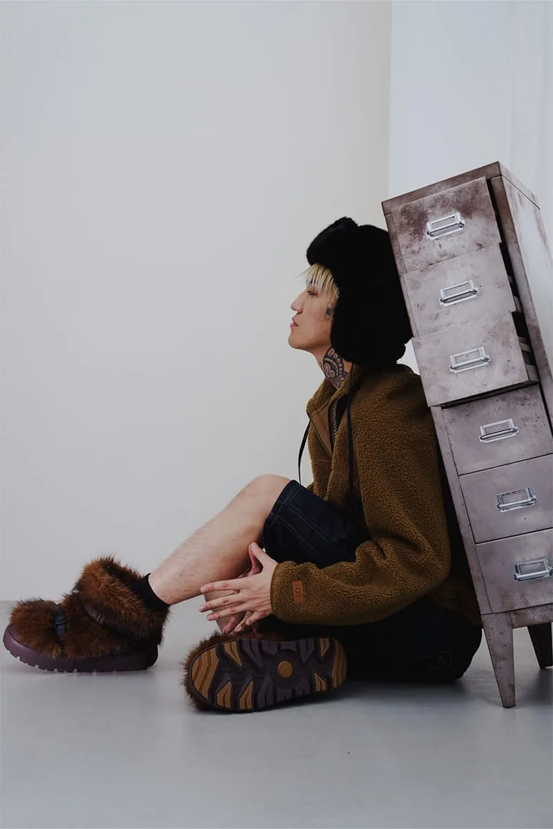 Seated on-foot shot of model in brown fur boots, one showing outsole, beside a metal filing cabinet, Heritage Utility Grizz