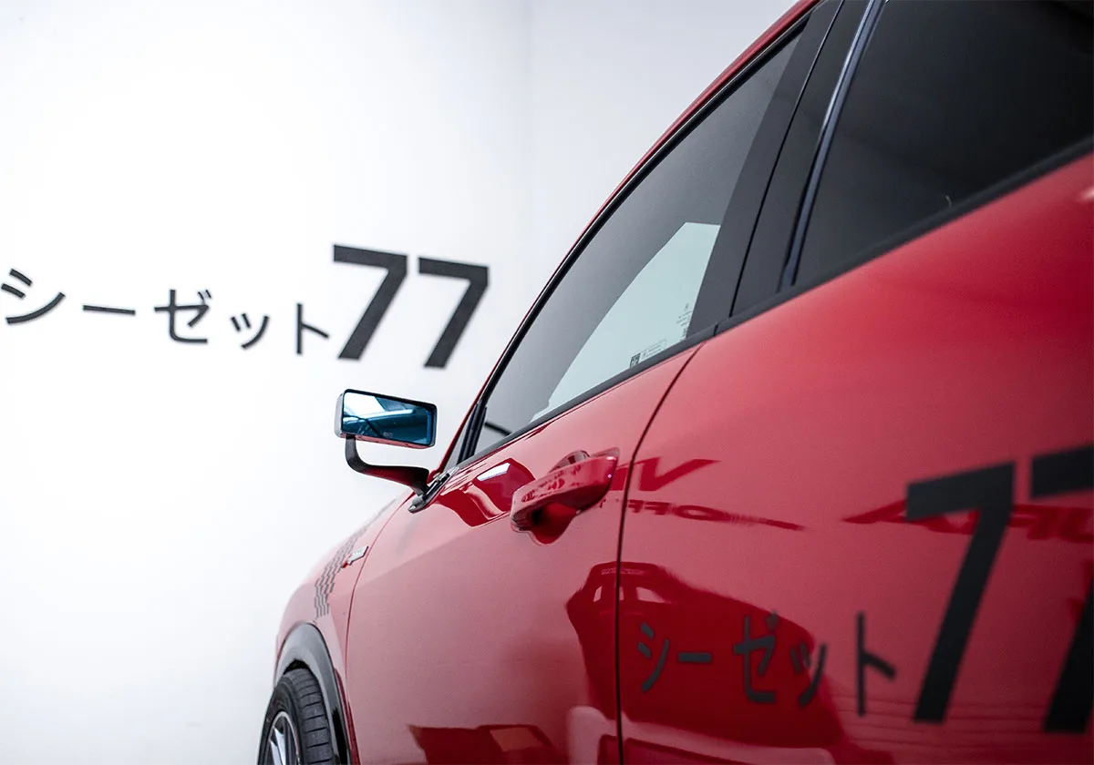 Red car side with mirror and door handle against white wall with Japanese text and 77, x Coco Zurita