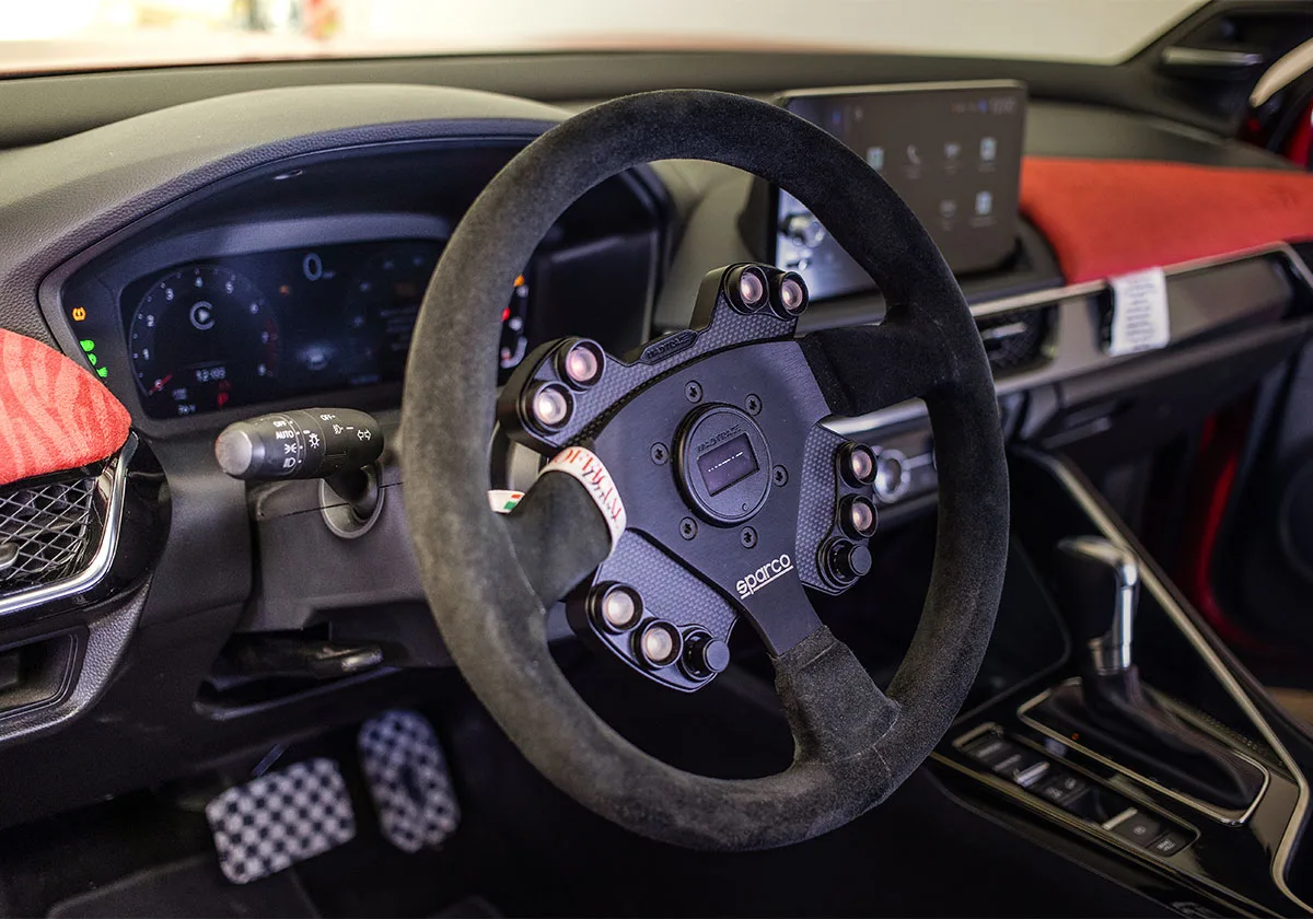 Interior view of black suede steering wheel with Sparco hub, red zebra dash trim, checkered pedals, x Coco Zurita