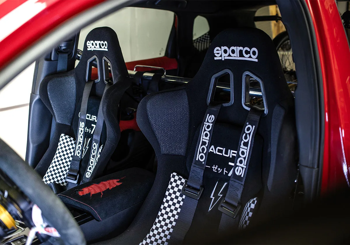 Two black Sparco racing seats with checkerboard harness pads, ACURA embroidery, red car body, x Coco Zurita