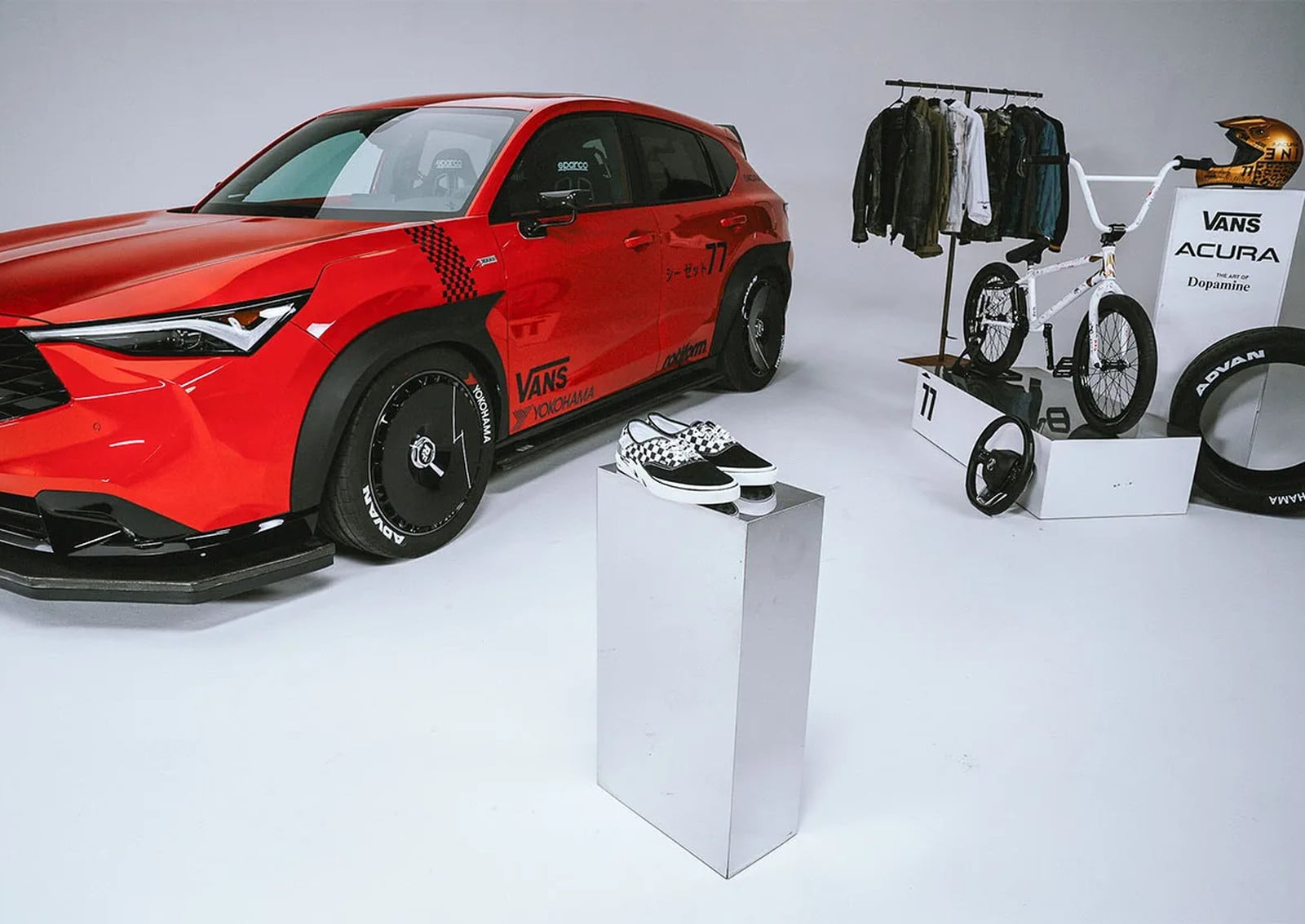 Shoes on mirrored plinth beside red race-styled car, bike and clothing rack display, x Coco Zurita