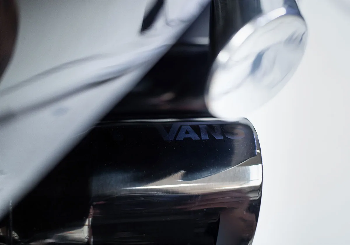 Close-up of reflective black chrome surface with VANS lettering, blurred white shape, x Coco Zurita