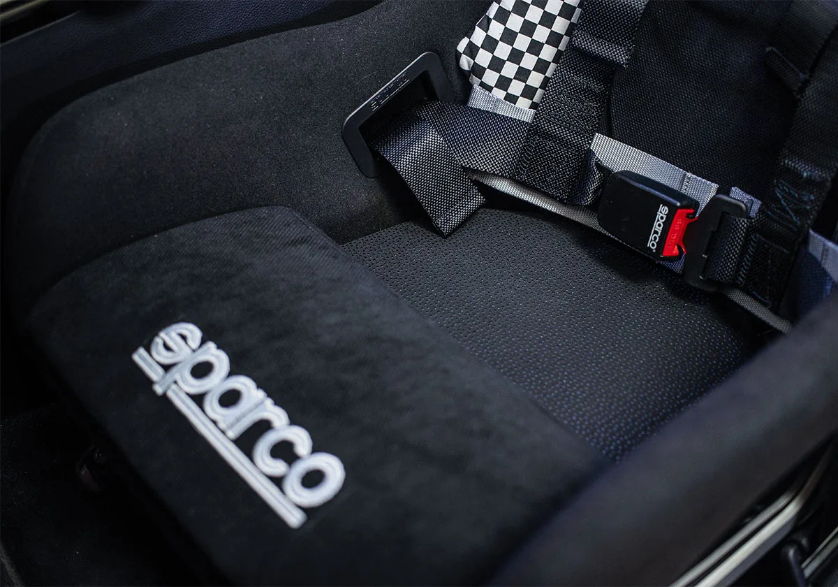 Detail of black racing seat with white Sparco logo and checkerboard harness pad, x Coco Zurita