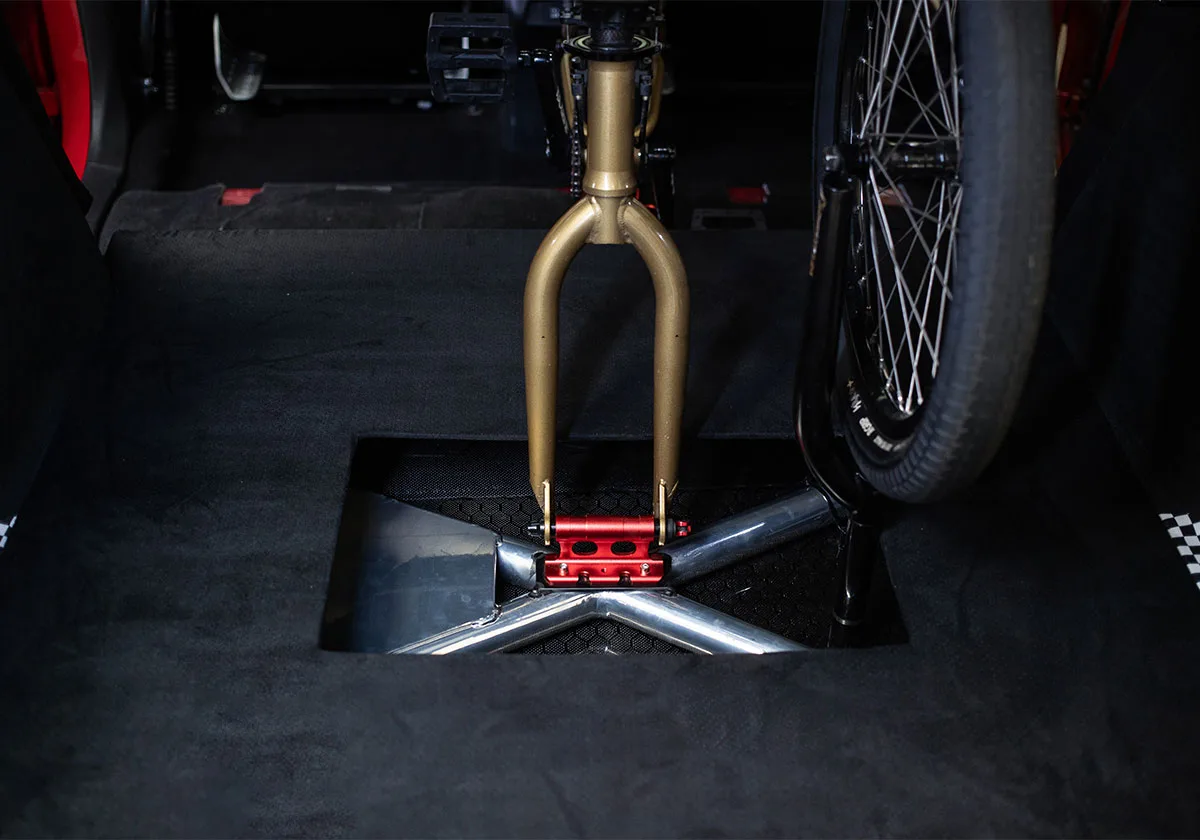 Gold bike fork mounted in a recess in black carpeted car floor, wheel at right, x Coco Zurita