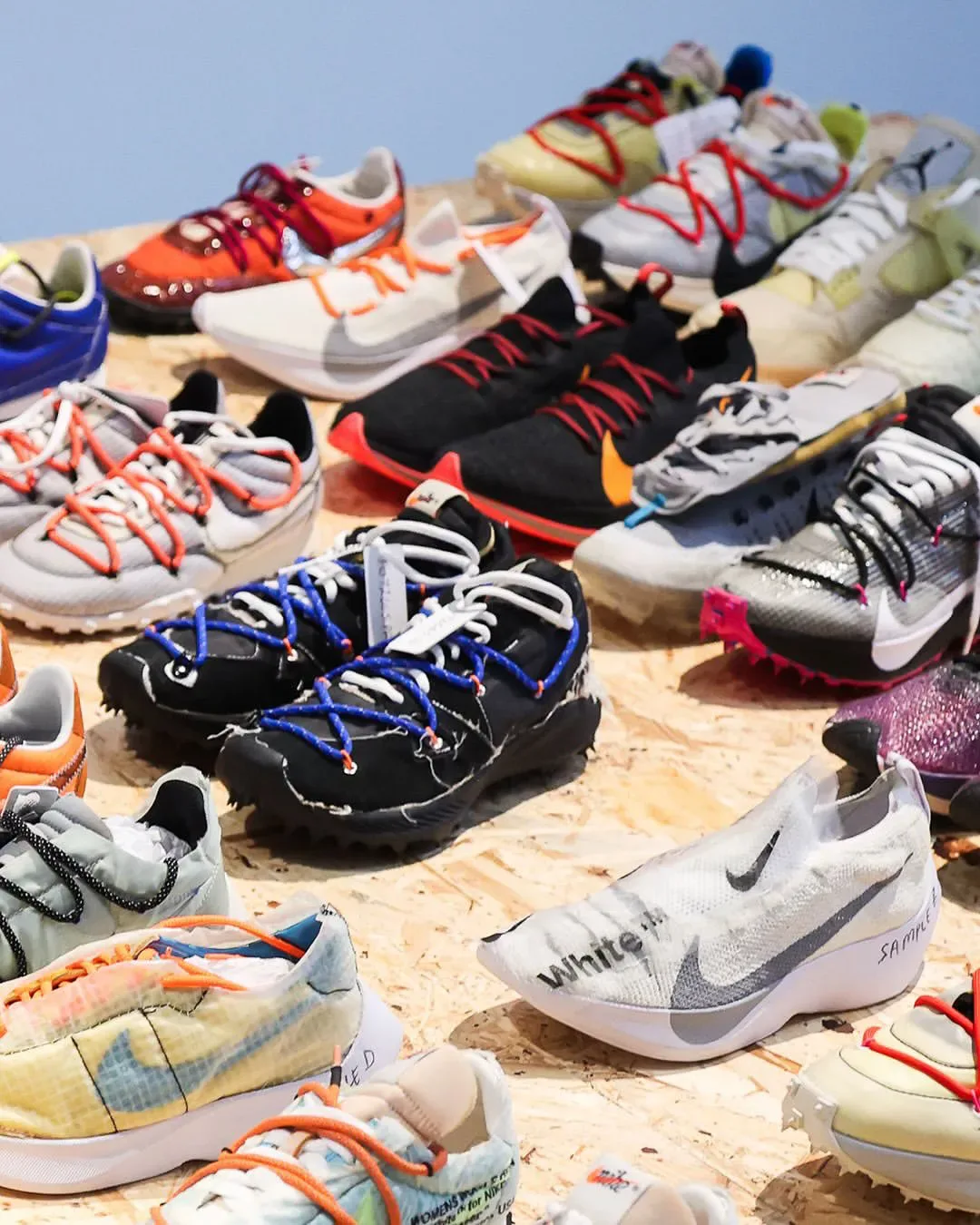 Top-down angle of many sneakers on plywood, black pair with blue laces, white pair marked "White", Abloh: The Codes
