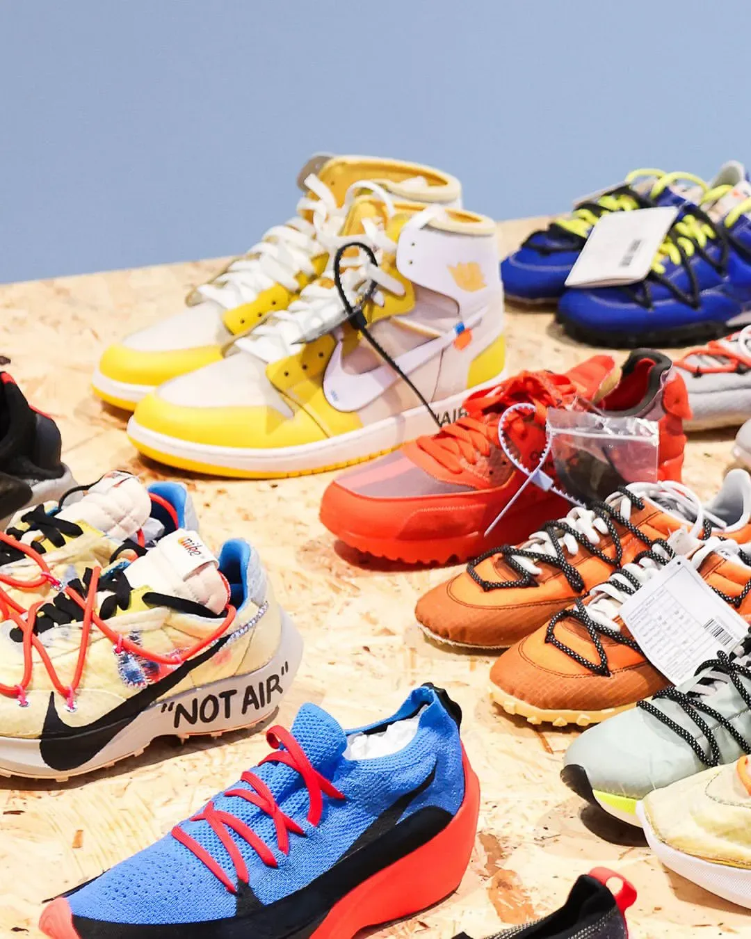 Many sneakers on a plywood table, yellow high-top, orange pair, blue knit and "NOT AIR" text, Abloh: The Codes